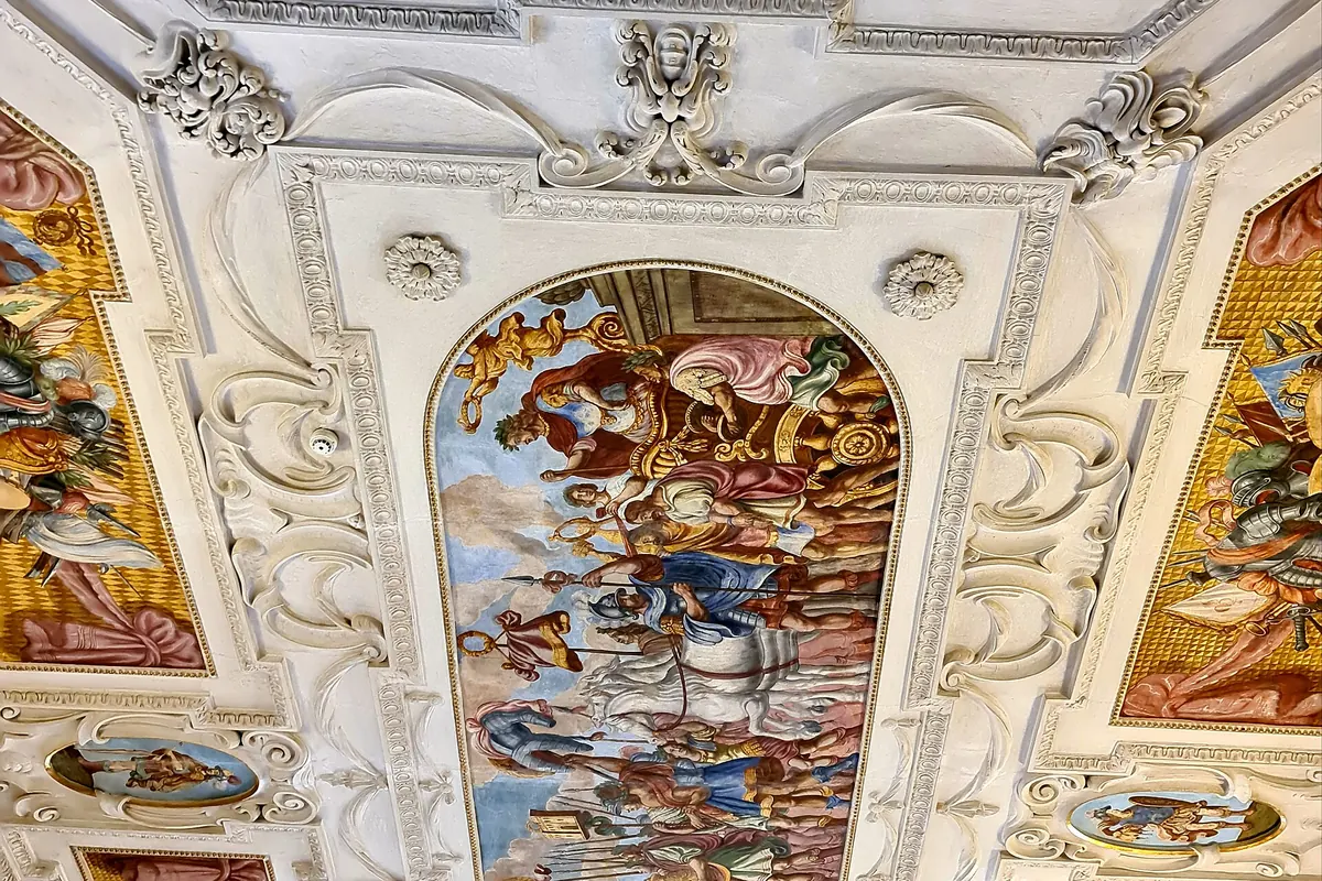Eine bemalte und stuckierte Decke im Lobkowicz-Palast mit einem großen zentralen Fresko, eingerahmt von weißem Stuckornament.