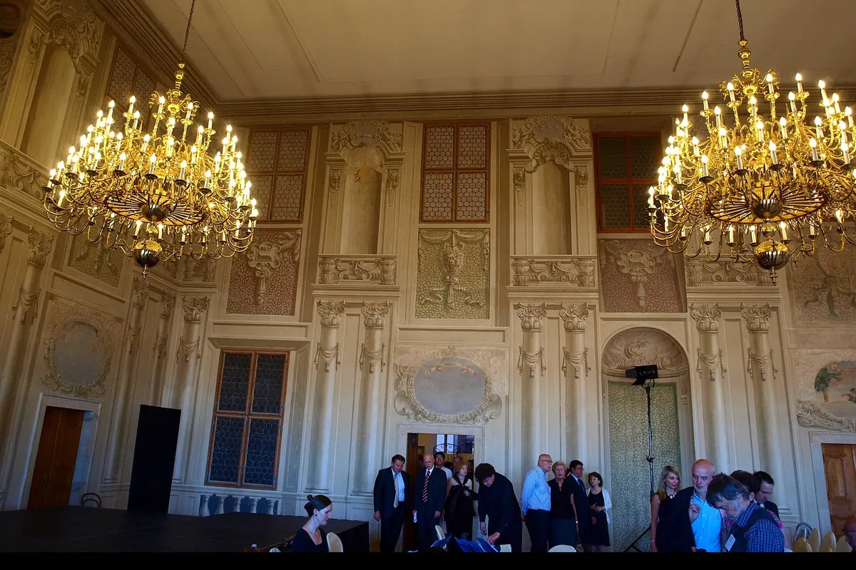 Ein Saal des Lobkowicz-Palastes mit zwei vergoldeten Kronleuchtern, bemalten Wandpaneelen und Stuhlreihen für eine Aufführung.