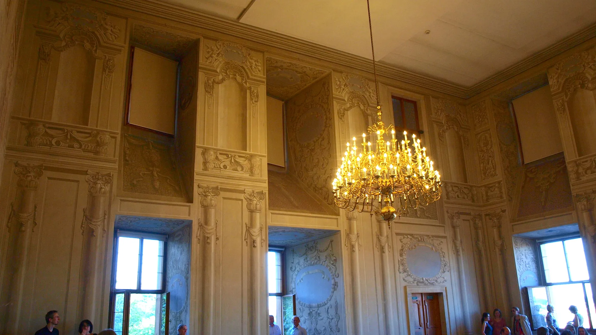 Ein hoher Saal im Lobkowicz-Palast mit bemalten Architekturpanelen an den Wänden, einem erleuchteten vergoldeten Kronleuchter rechts und hohen Fenstern darunter.